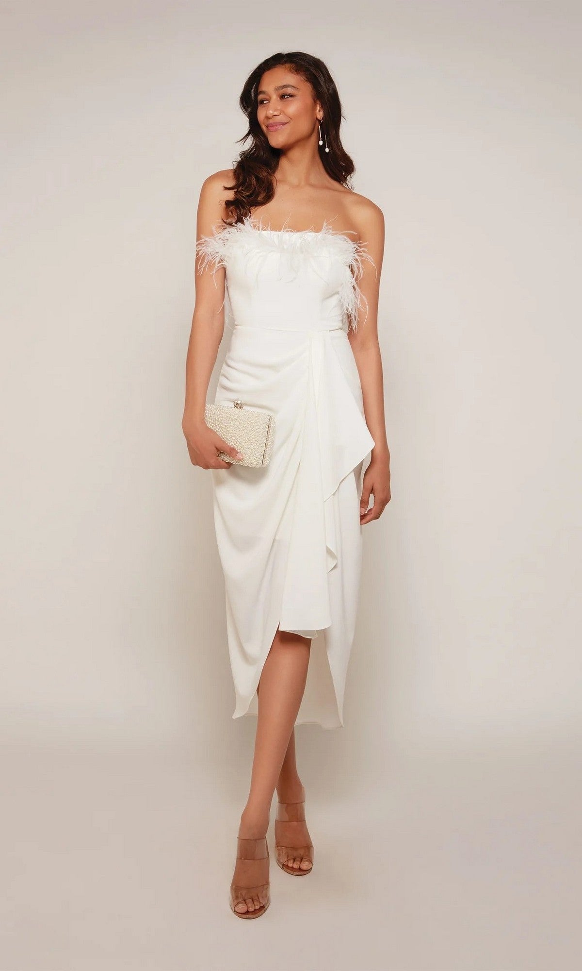 A woman poses against a plain light background wearing the Alyce 70034 knee length semi-formal strapless white dress with feather accents, complemented by a beige clutch and nude heels.